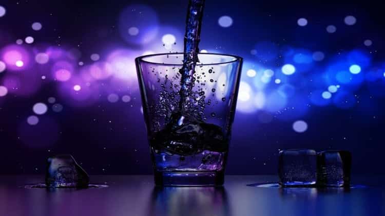 A stream of water pours into a clear glass against a vibrant blue, purple and white background