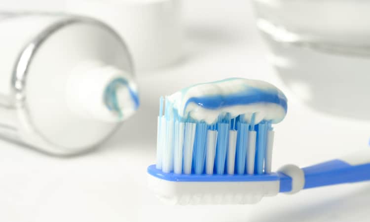 Closeup of a white and blue soft-bristled toothbrush with a stream of toothpaste next to a white tube of toothpaste and a water glass