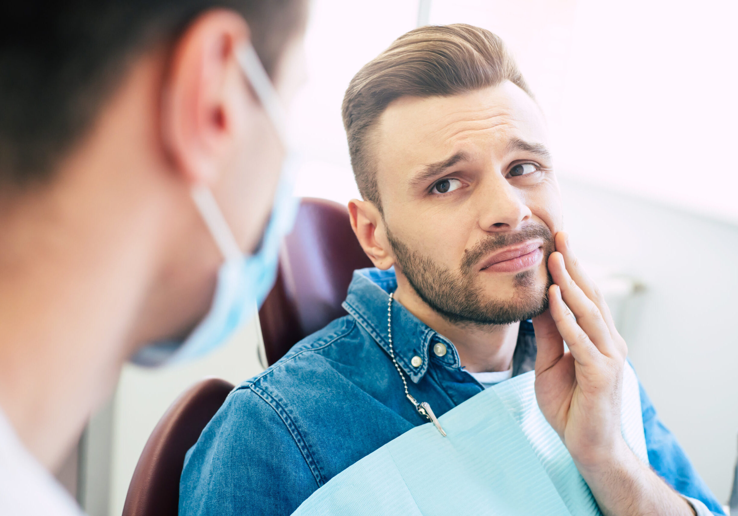 Symptoms of gums pain. A man with a worried face is holding his hand on his cheek because of irritating pain in front of a dentist who is going to give a patient a treatment. Symptoms of gums pain. A man with a worried face is holding his hand on his cheek because of irritating pain in front of a dentist who is going to give a patient a treatment.
