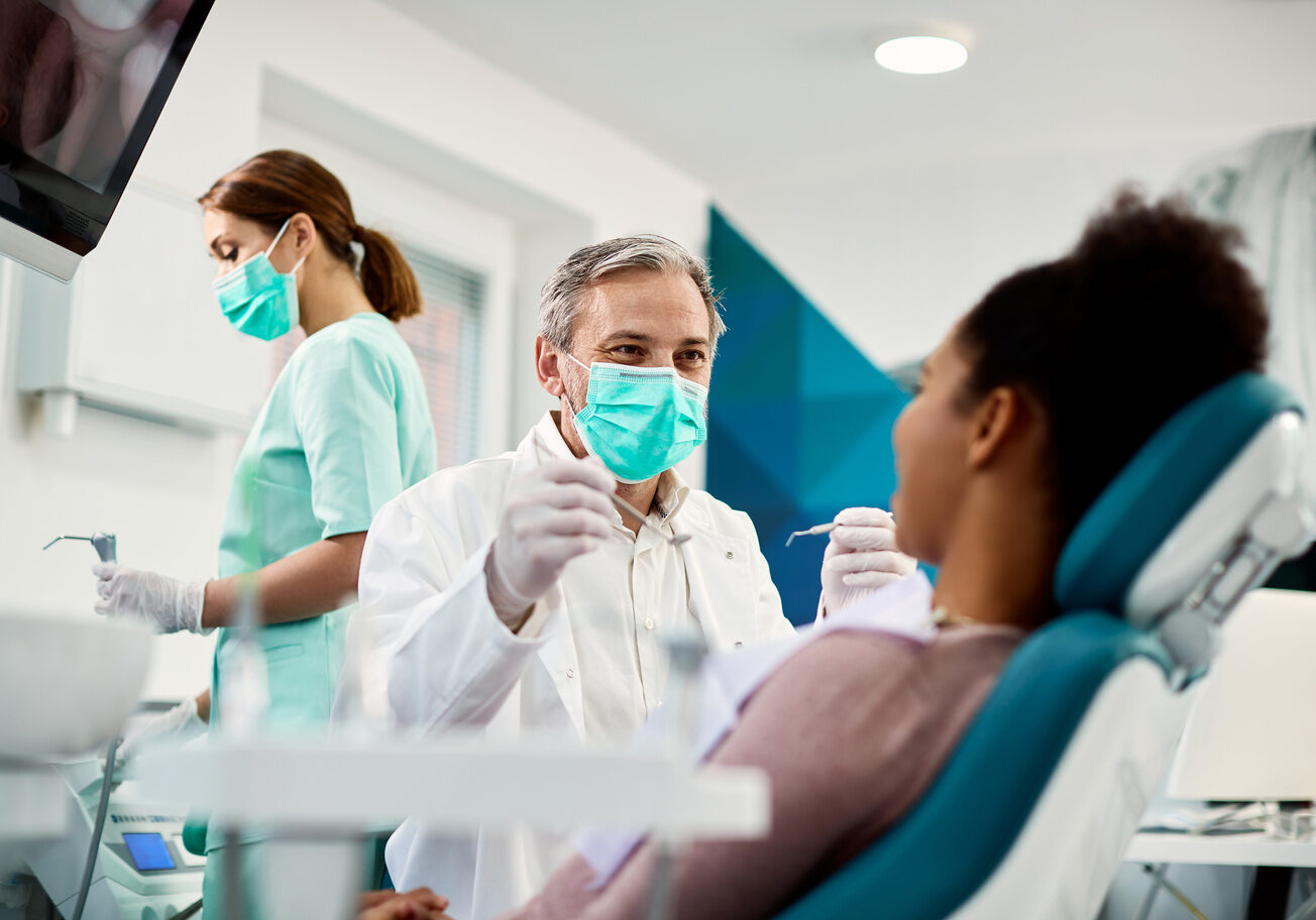 Smiling dentist with face mask talking to Black woman during dental procedure at dental clinic. Smiling dentist communicating with African American woman while checking her teeth during dental procedure at dentist's office.